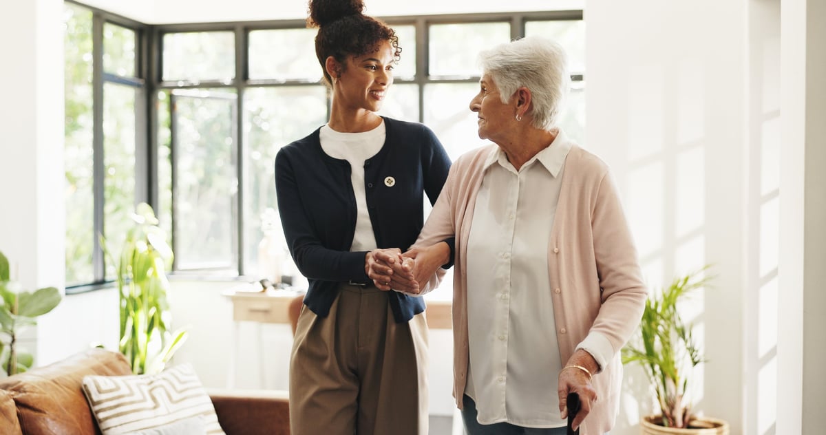 Caregiver supporting an African American senior woman with a walking stick in her home